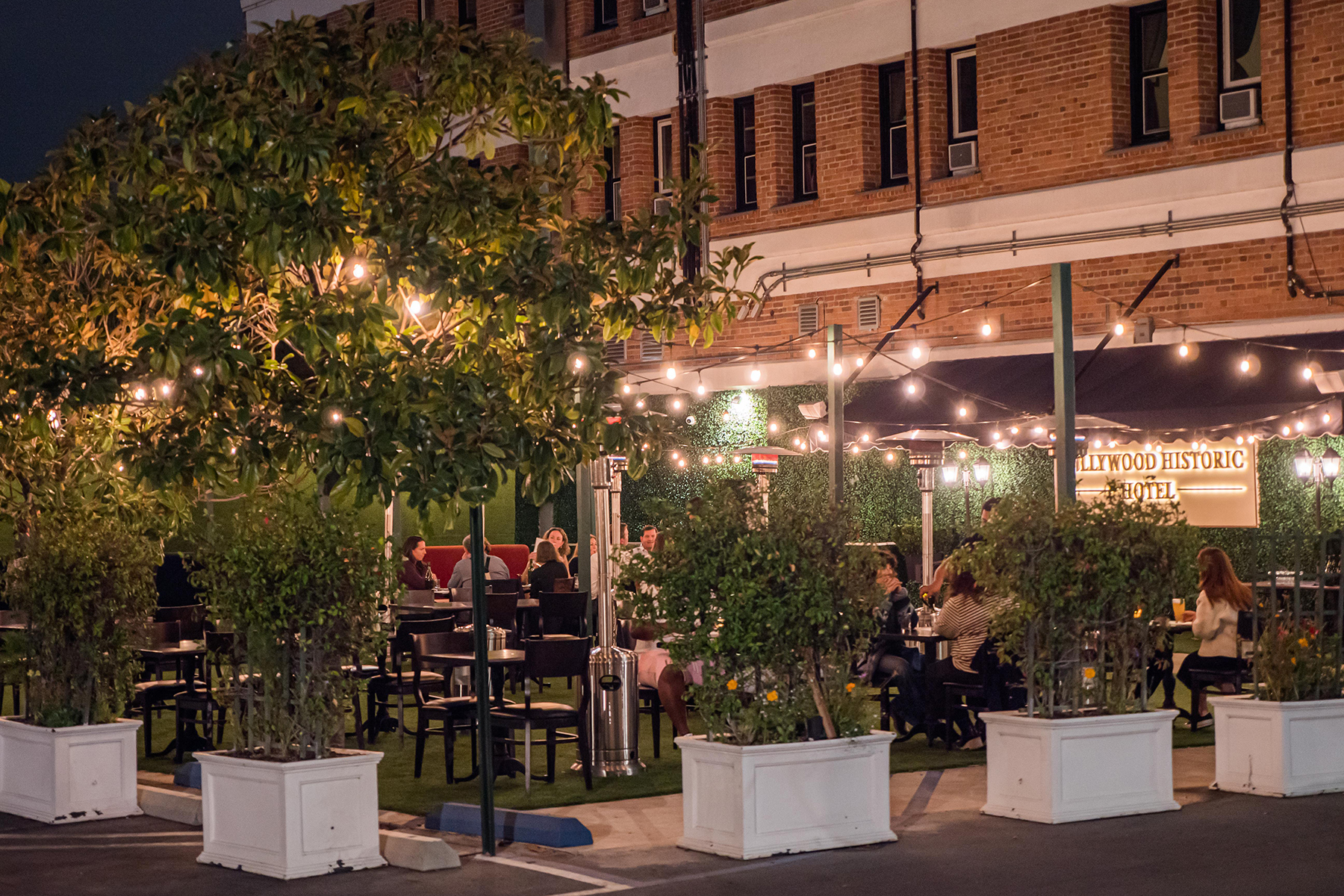 A nighttime view of a brick building with outdoor seating and string lights, featuring a sign that reads 'Hollywood Historic Hotel'.