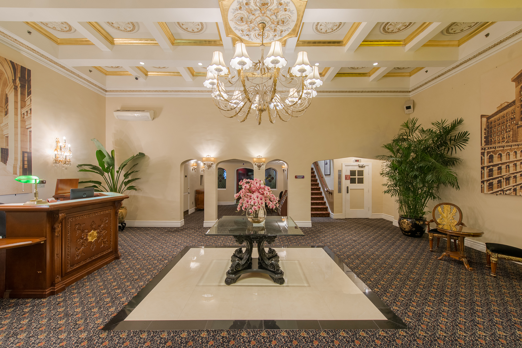 A hotel lobby with a chandelier, reception desk, plants, and staircase.