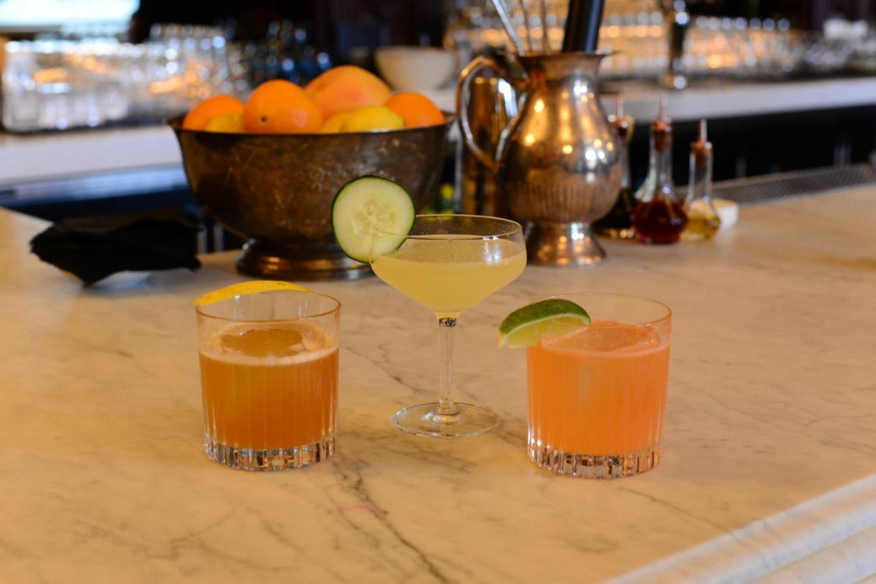 three cocktails on a marble bar counter with a bowl of fruits in the background