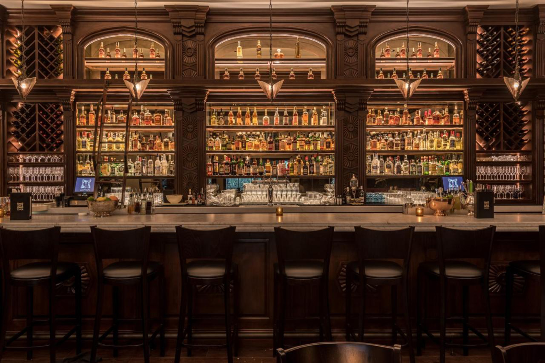 A dimly lit bar with a long counter and several stools, behind which are shelves stocked with various bottles and glasses.